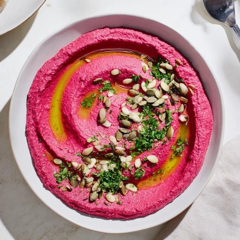 A bowl of creamy Beet Hummus topped with olive oil, seeds, and fresh parsley, with pita and veggies.